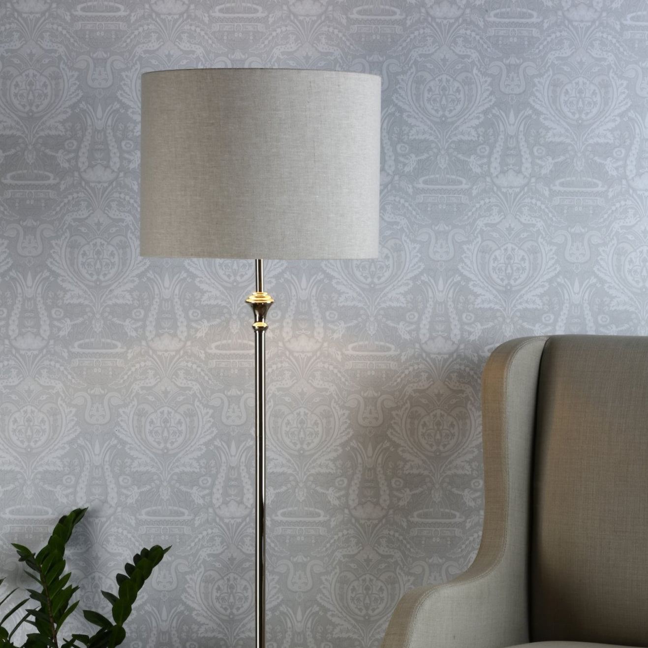 A floor lamp with a neutral shade and chrome stand, illuminating a patterned wall and a chair.