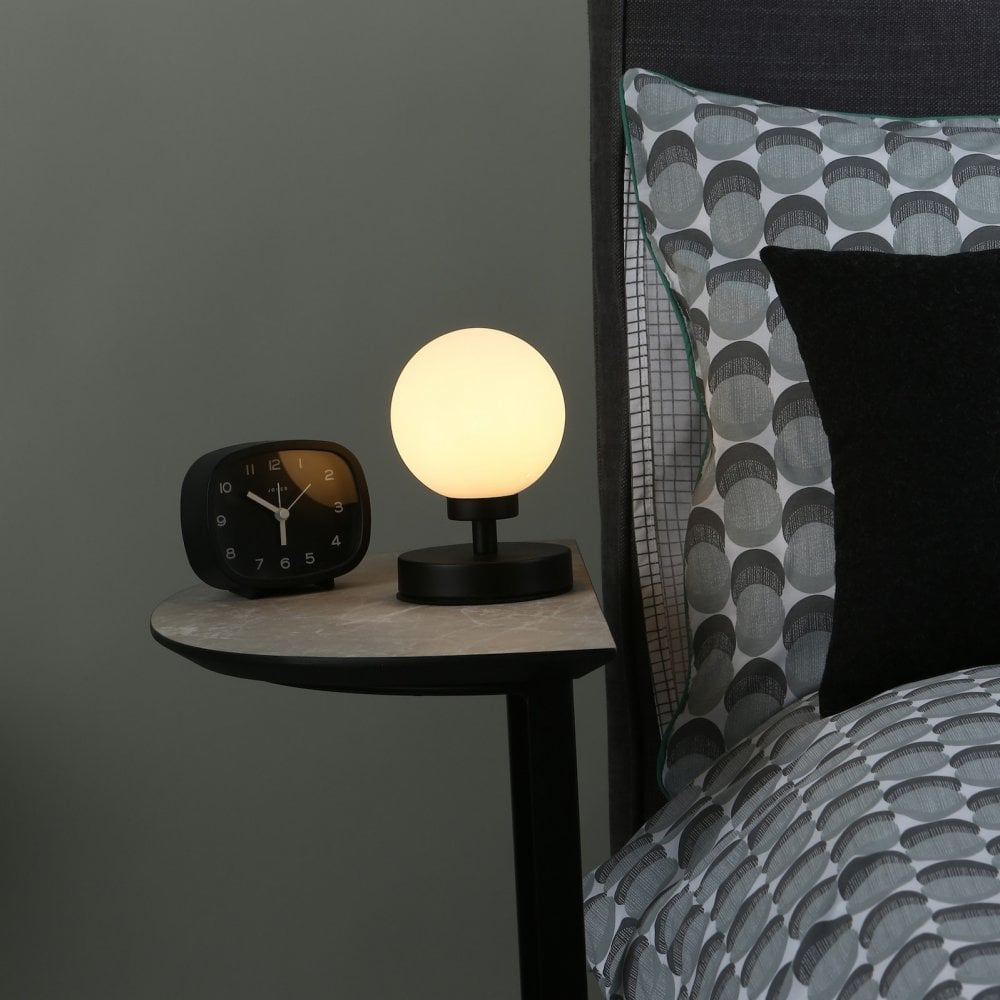 A small spherical table lamp illuminating a bedside table next to a patterned duvet.