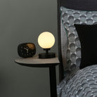 A small spherical table lamp illuminating a bedside table next to a patterned duvet.