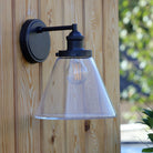 Vintage outdoor wall light with a black fixture and clear conical glass shade, unlit on wood.