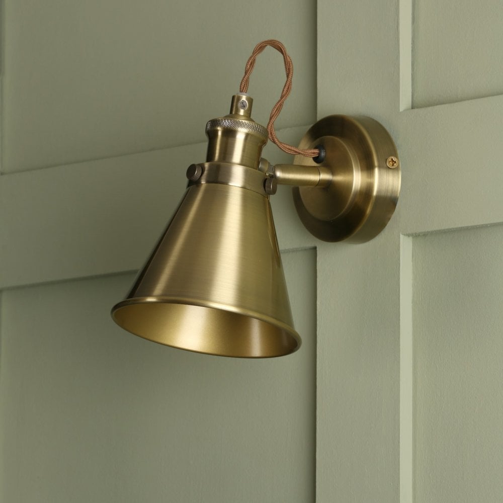 A close-up of an industrial brass cone wall light with a braided cable on a green wall.