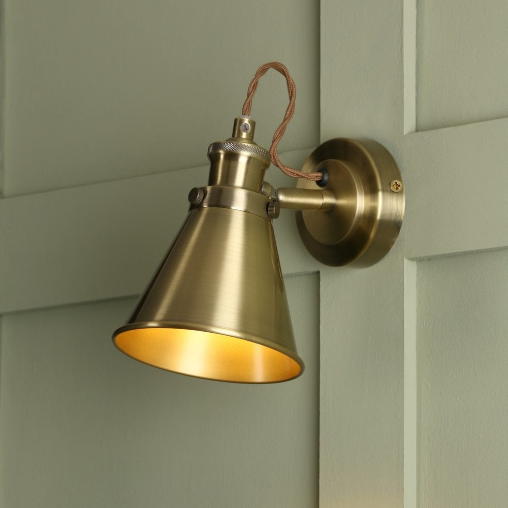 An industrial brass cone wall light with a braided cable on a paneled green wall.