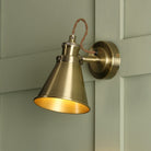 An industrial brass cone wall light with a braided cable on a paneled green wall.