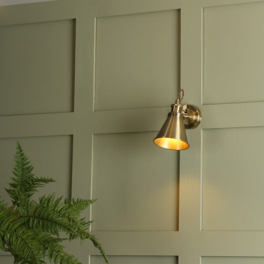 An industrial brass cone wall light with warm light on a paneled green wall with a plant.