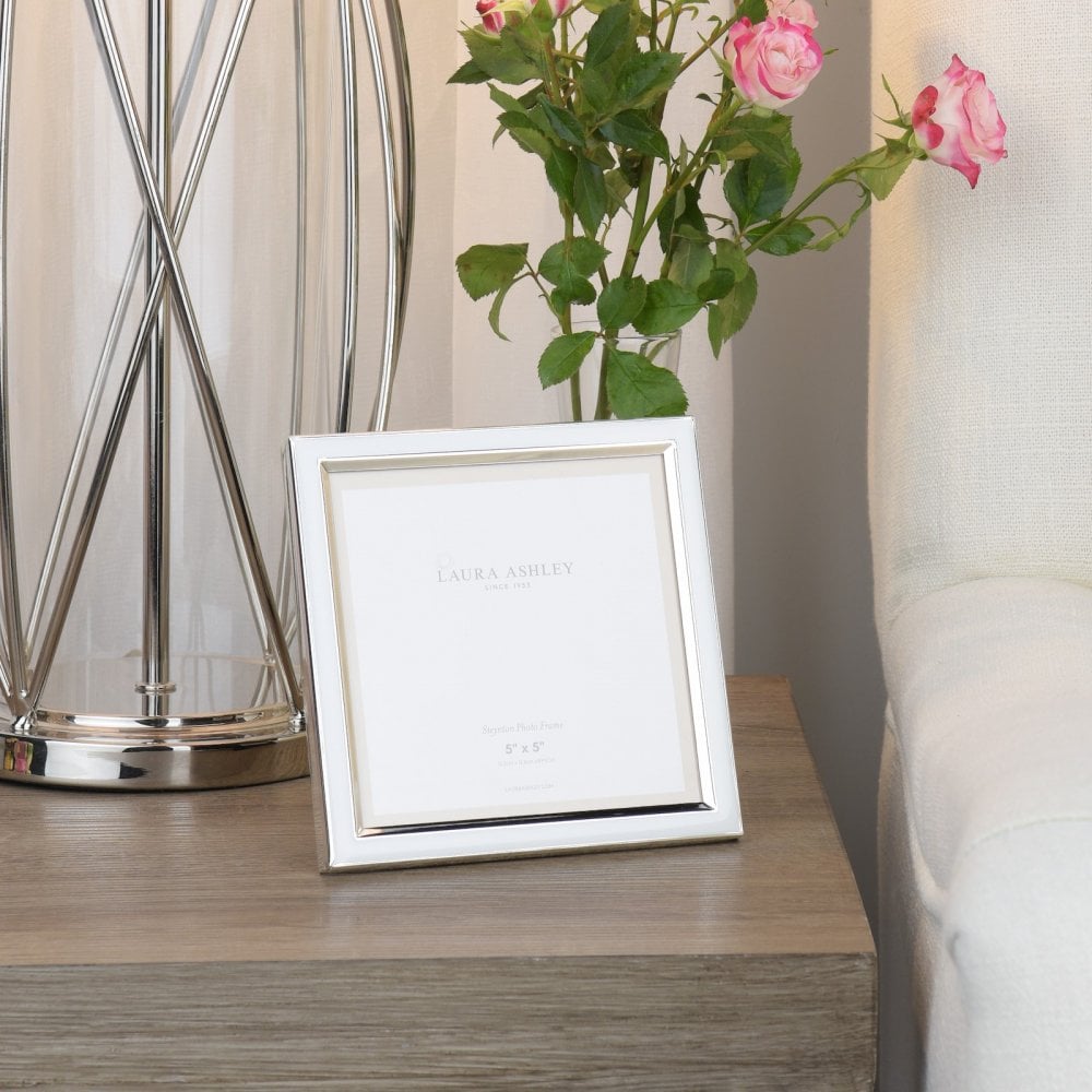A white photo frame sits on a wooden bedside table, next to a silver lamp and pink roses.