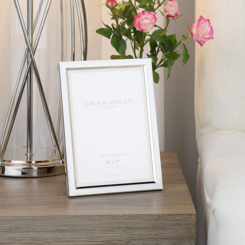 Silver photo frame on a wooden bedside table with a lamp and pink flowers.