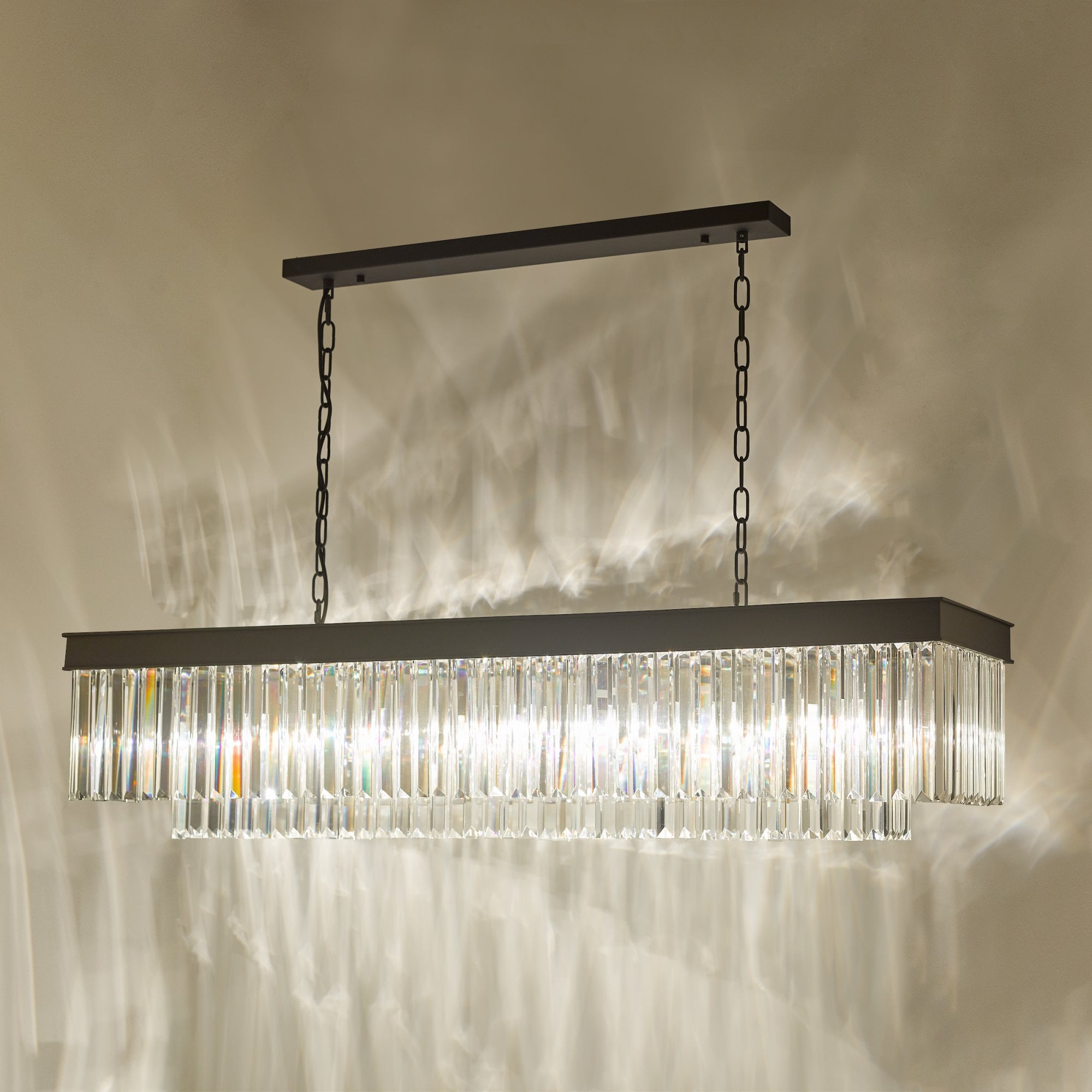 A rectangular chandelier with a black frame and crystal rods illuminating a wall.