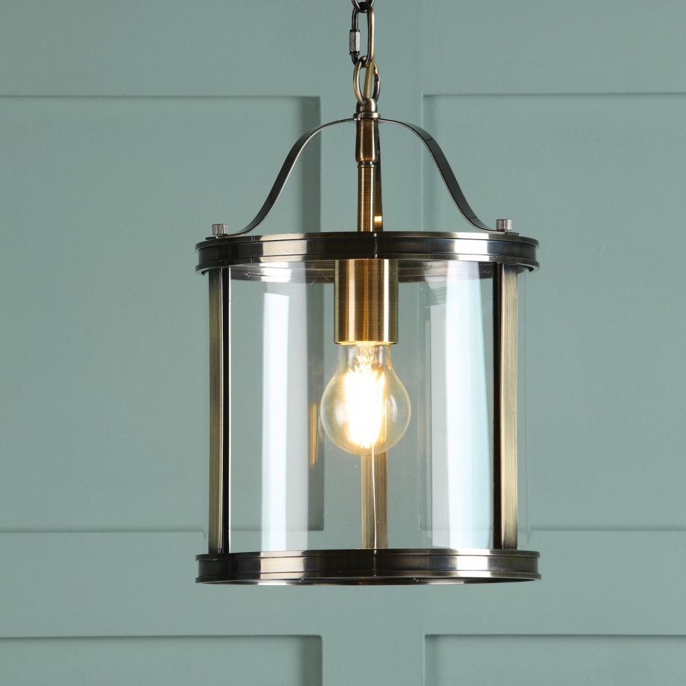 A vintage style pendant light with a clear glass shade and antique brass finish against a teal wall.