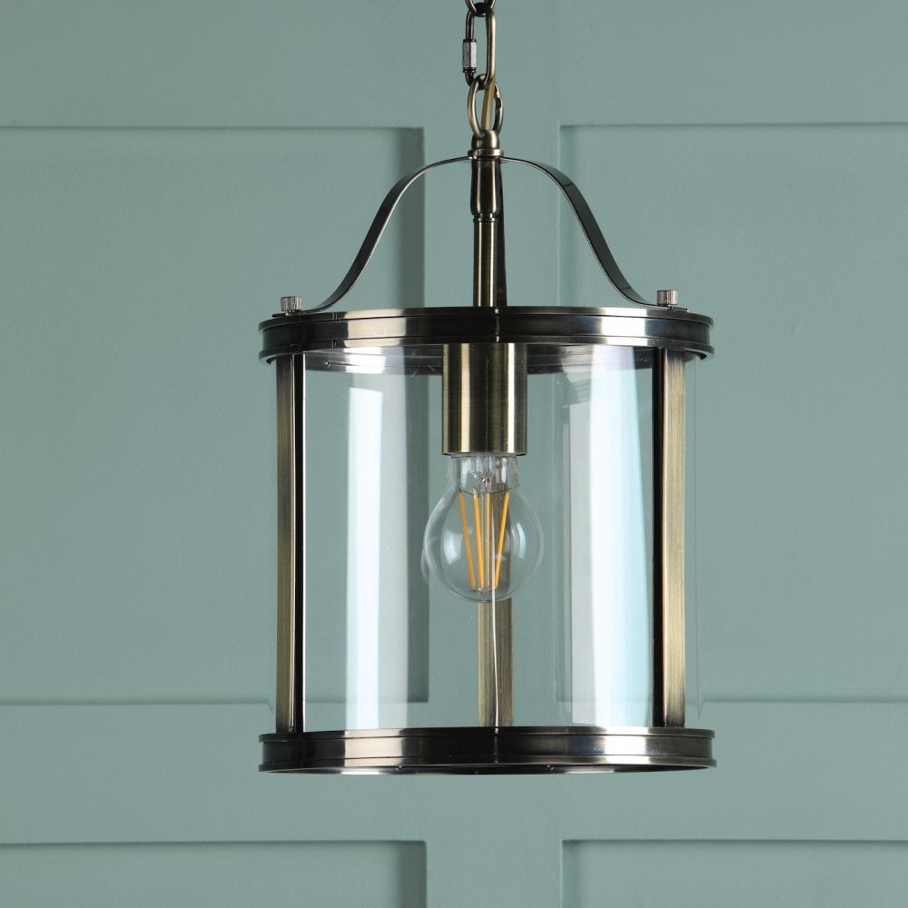 A vintage style pendant light with a clear glass shade and antique brass finish against a teal wall.