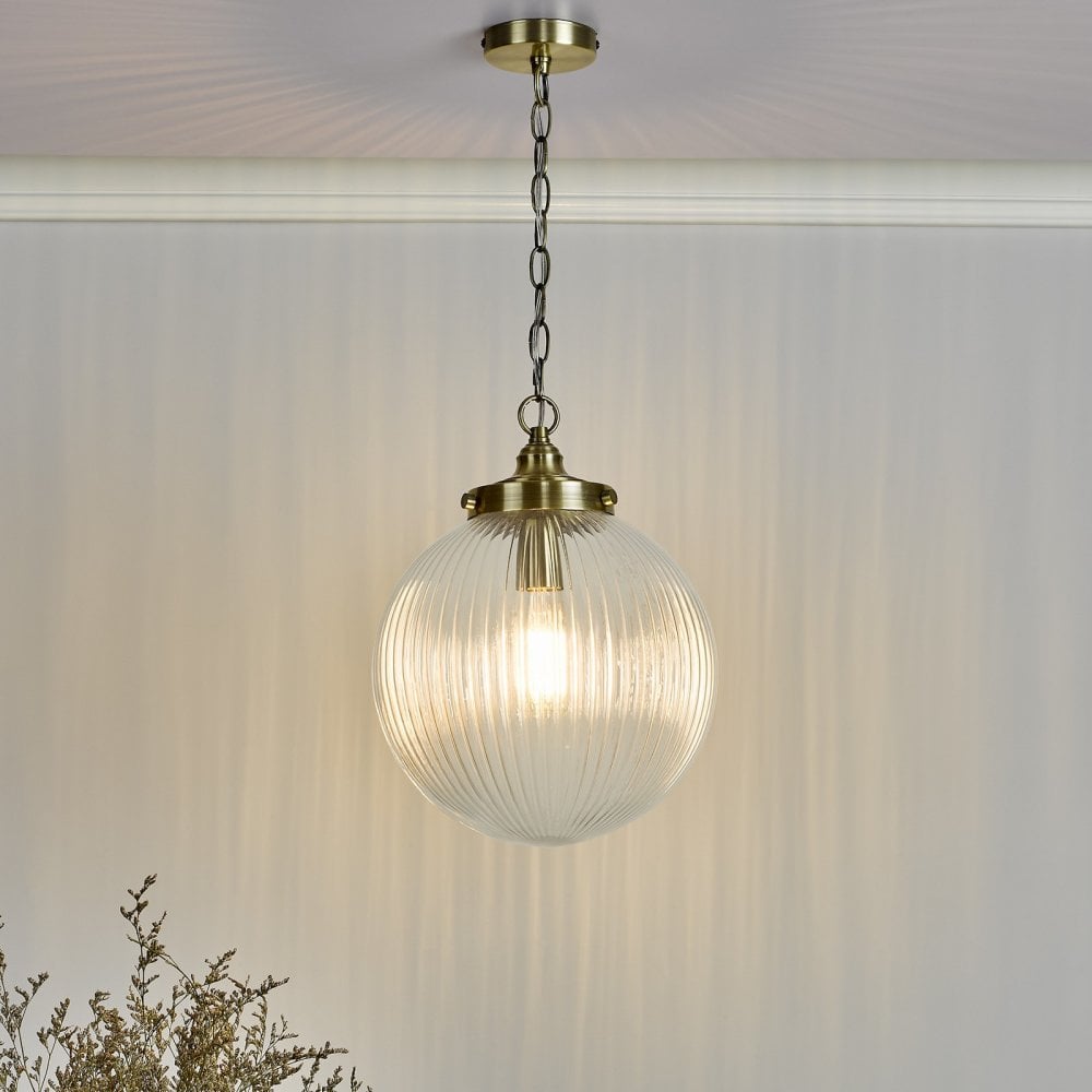 An illuminated ribbed glass globe pendant light with brass accents, hanging in a room with light walls.