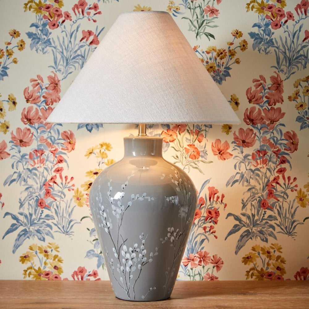 A white tapered lampshade on a grey base with white flowers in front of floral wallpaper.