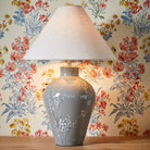 A white tapered lampshade on a grey base with white flowers in front of floral wallpaper.