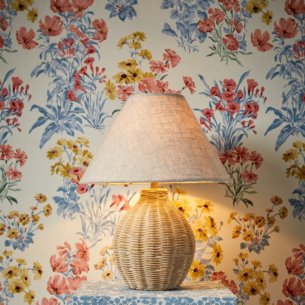 A wicker table lamp with a soft glow sits on a patterned blue table against floral wallpaper.