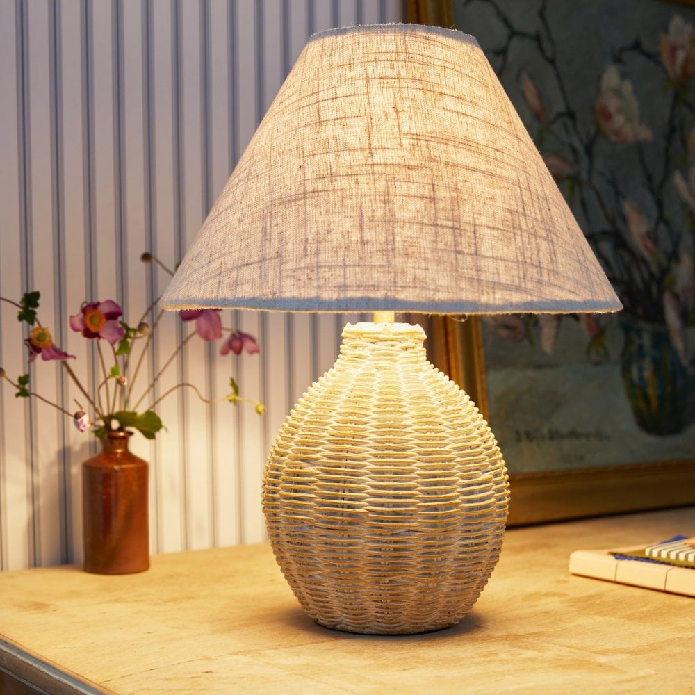 A wicker table lamp illuminates a wooden desk with a small vase of flowers and a painting.