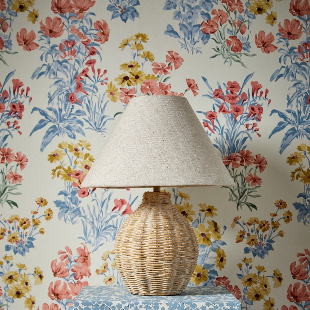 A wicker table lamp on a patterned blue table in front of vibrant floral wallpaper.
