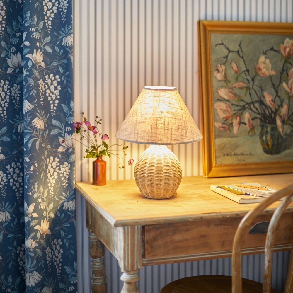 A wicker table lamp illuminates a desk next to a blue floral curtain and a framed painting.