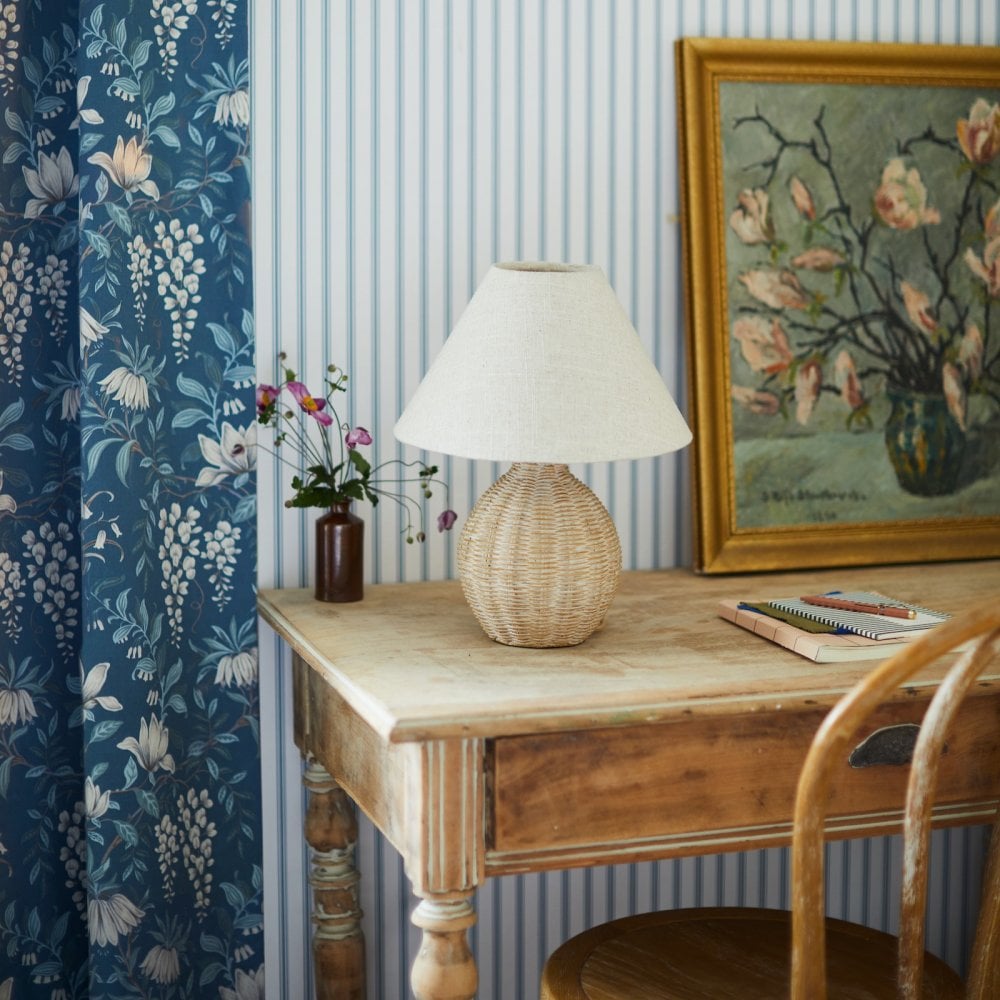 A wicker table lamp on a desk with a vase of flowers, painting, and blue floral curtain.