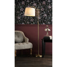 A brass floor lamp with a pleated shade next to an armchair and a table with flowers.