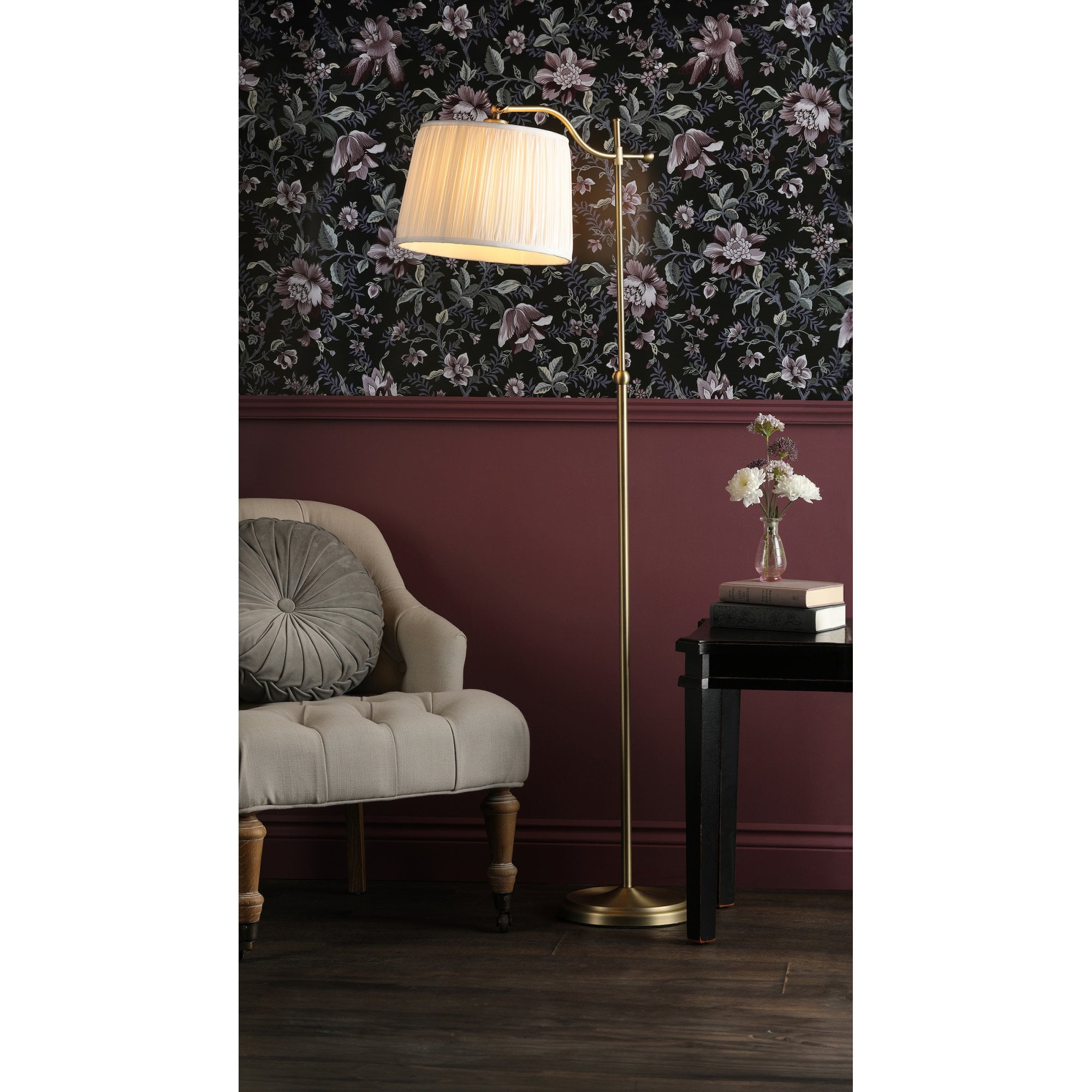 A brass floor lamp with a pleated shade next to an armchair and a table with flowers.