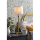 A brass table lamp with a pleated shade on a bedside table with floral wallpaper.