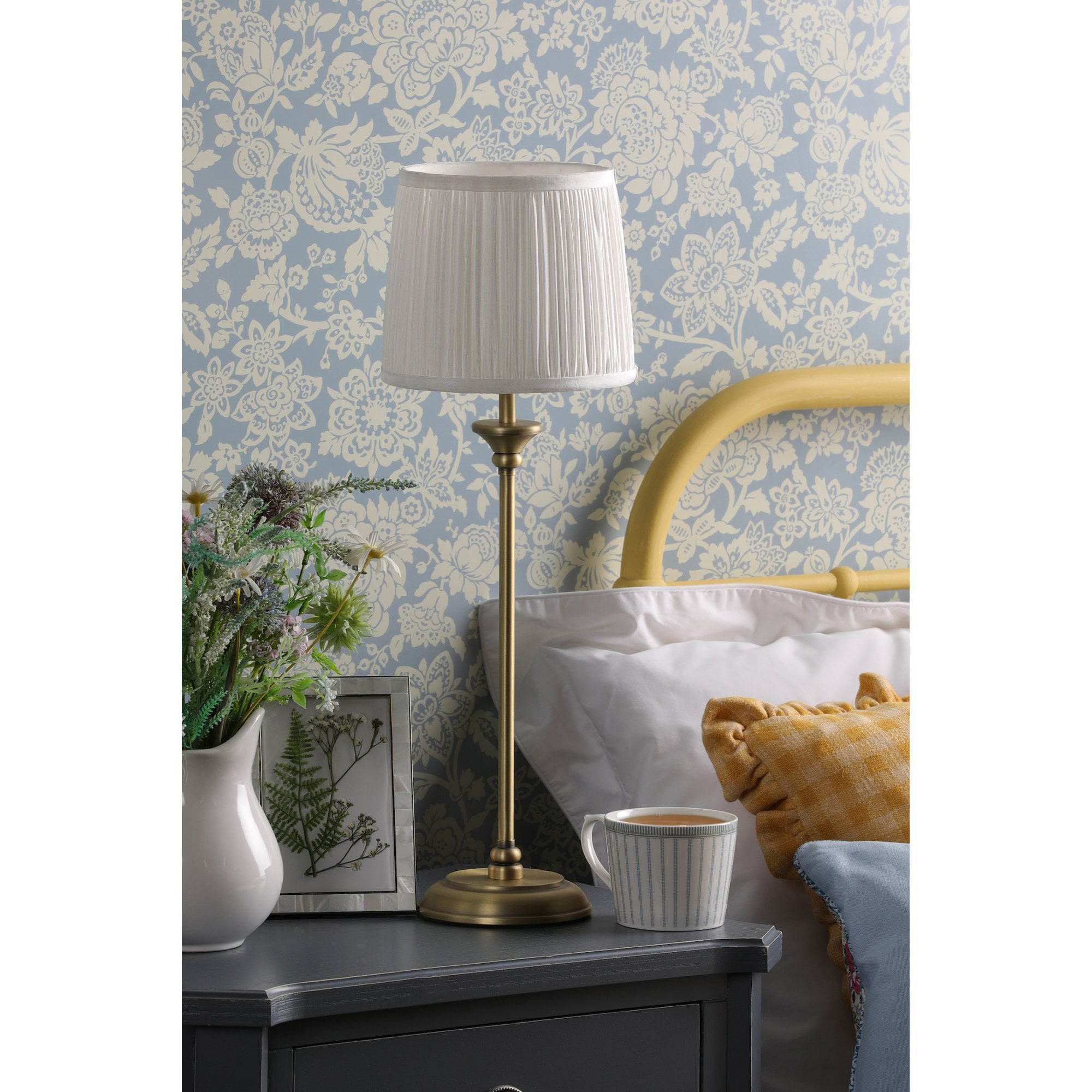 A tall brass table lamp with a white pleated shade on a bedside table.