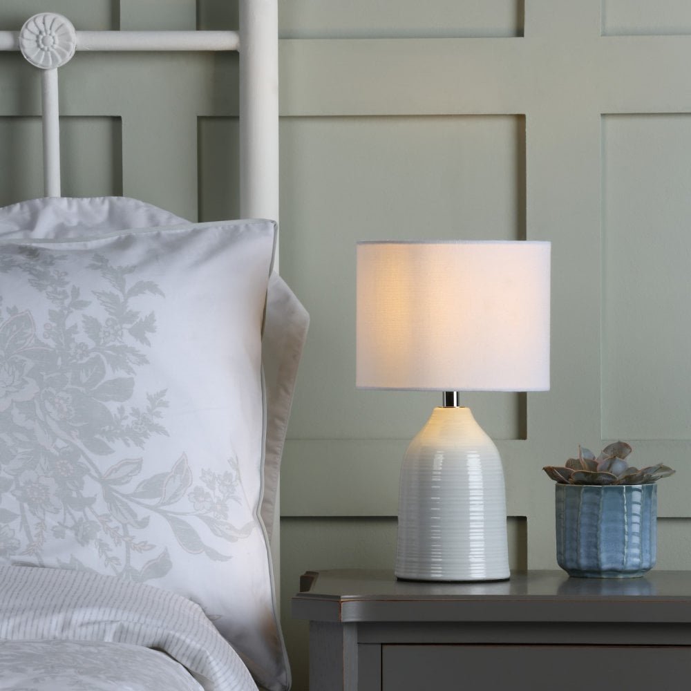 A white ceramic table lamp with a drum shade on a grey bedside table next to a bed.