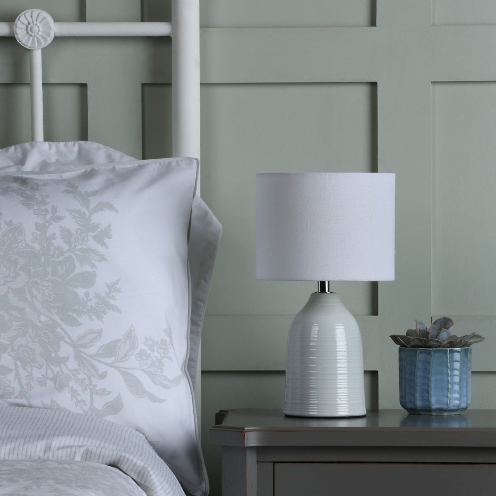 A white ceramic table lamp with a drum shade on a grey bedside table next to a bed.