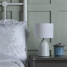 A white ceramic table lamp with a drum shade on a grey bedside table next to a bed.