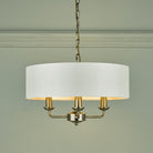 A brass three-arm pendant light with a white drum shade against a light green wall.