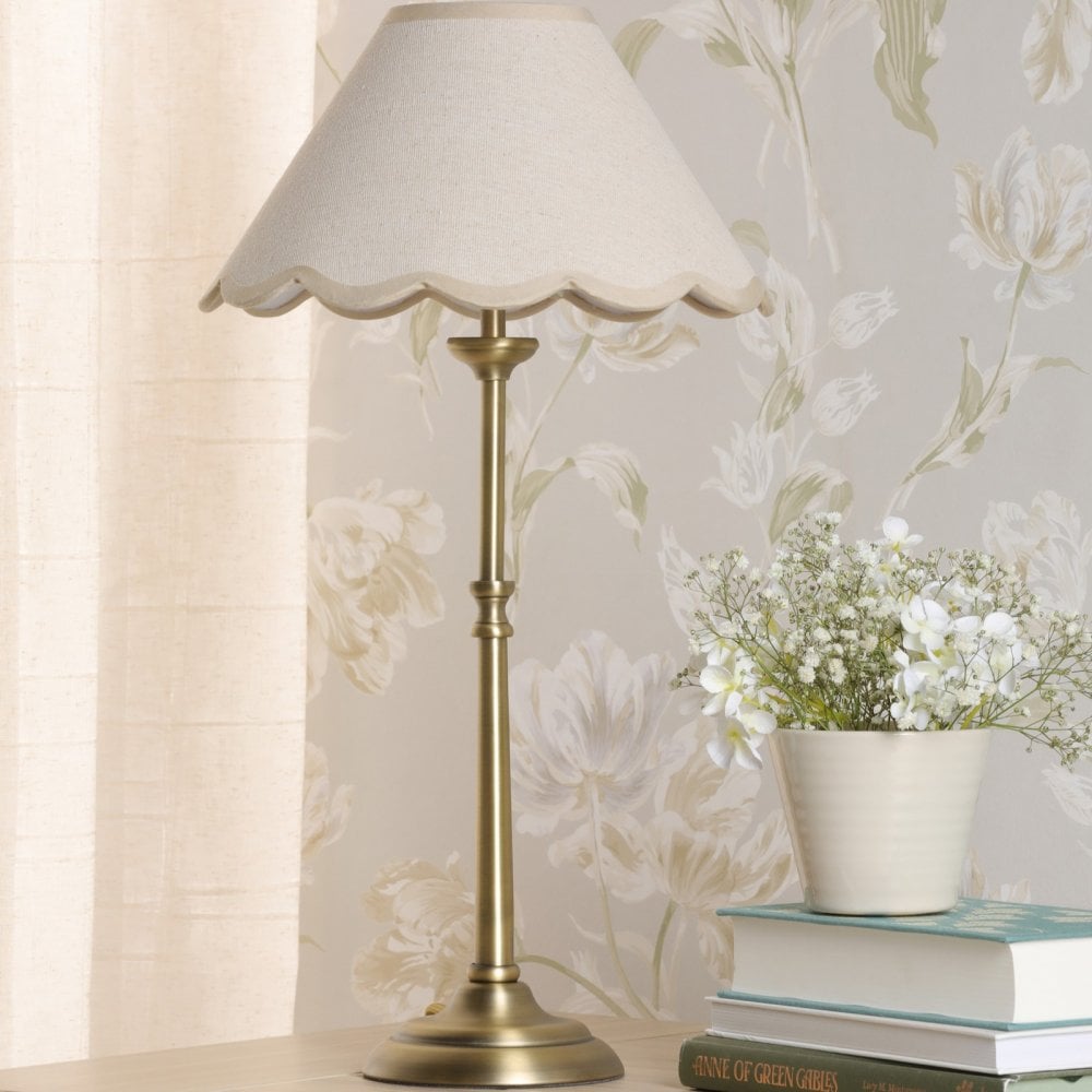 A brass table lamp with a scalloped cream shade, next to a vase of white flowers.