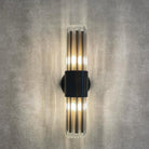 An illuminated black and clear glass rod wall sconce casting warm light on a grey wall.