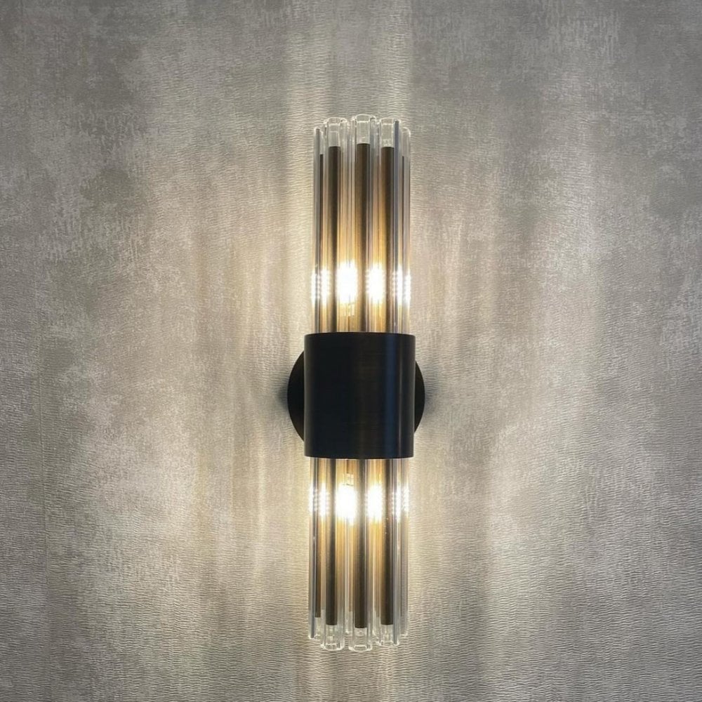 An illuminated black and clear glass rod wall sconce casting warm light on a grey wall.