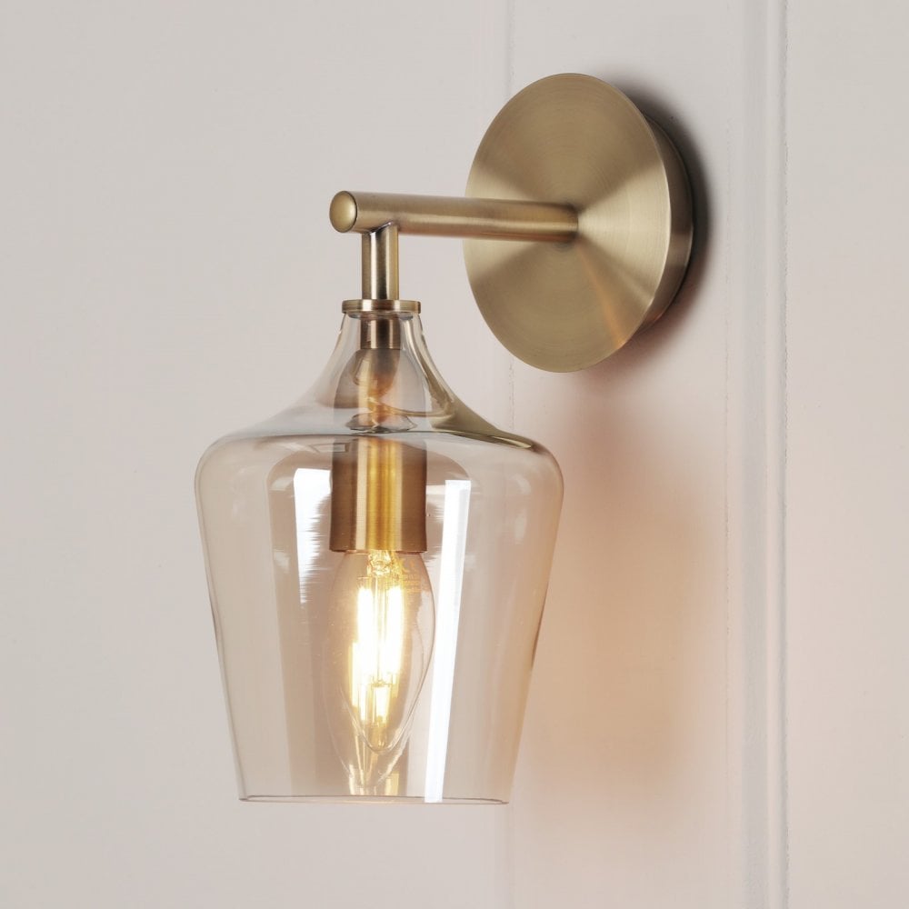 A brass wall light with a clear glass shade illuminating a warm glow on a white wall.