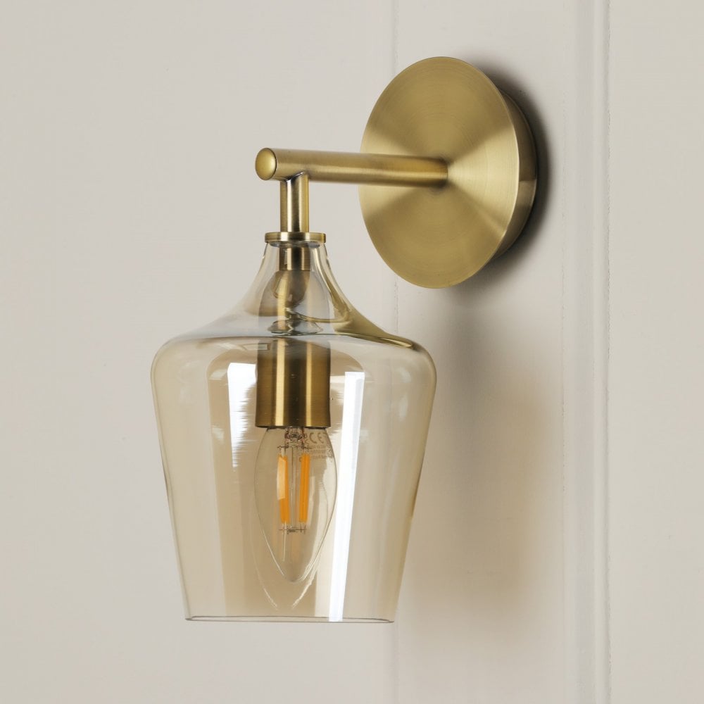 A brass wall light with a clear glass shade and a visible filament bulb on a white wall.