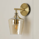 A brass wall light with a clear glass shade and a visible filament bulb on a white wall.