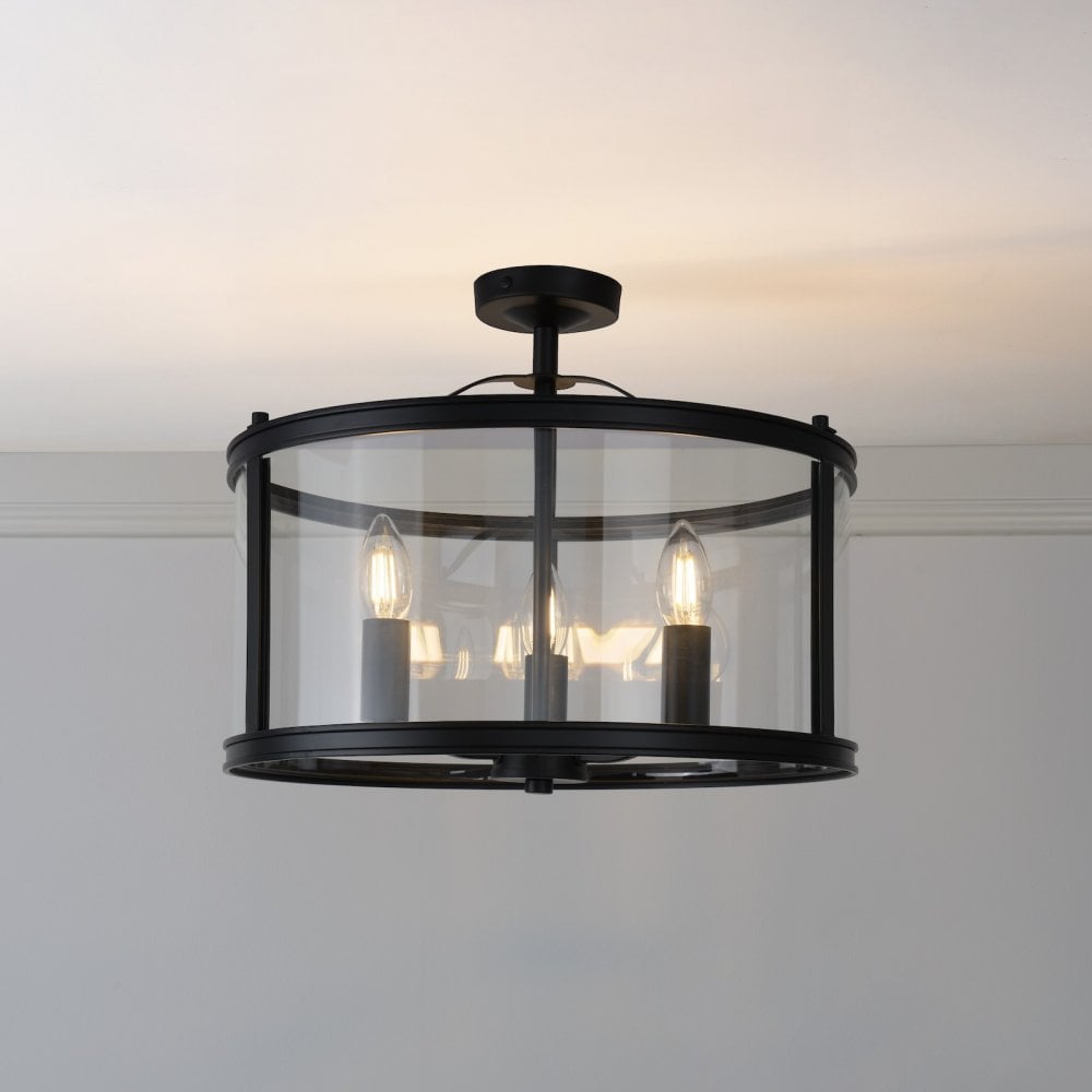 A sleek black semi-flush ceiling light with a clear glass drum shade, showcasing three bulbs.