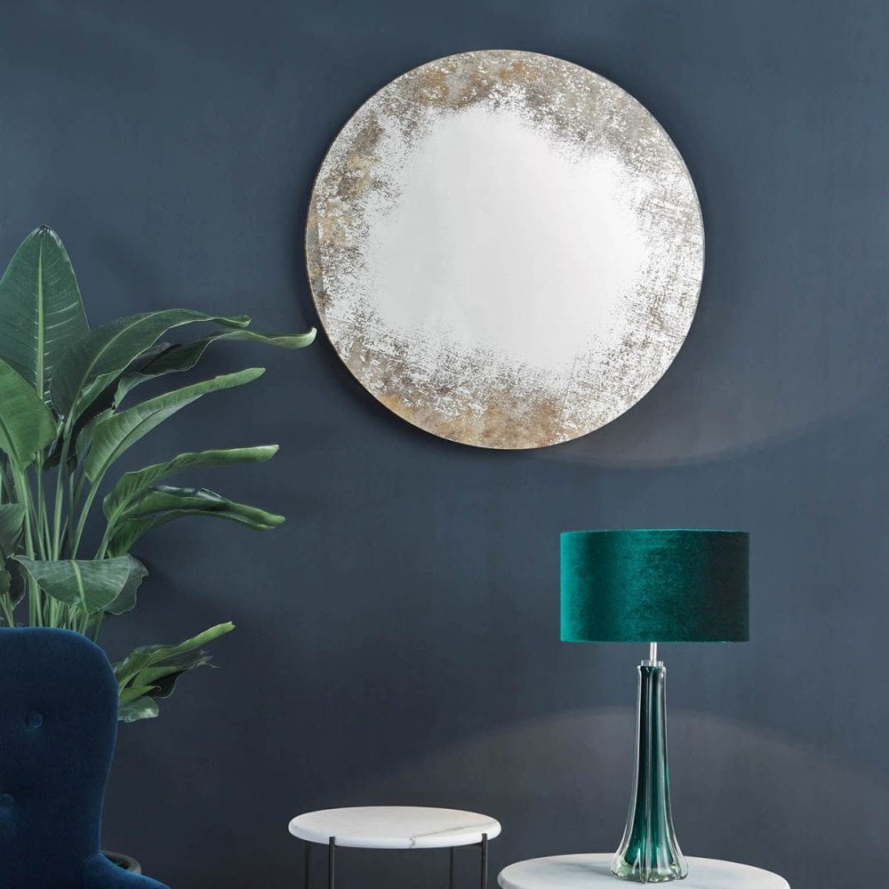A round distressed mirror hanging on a dark blue wall above a modern green lamp and plant.