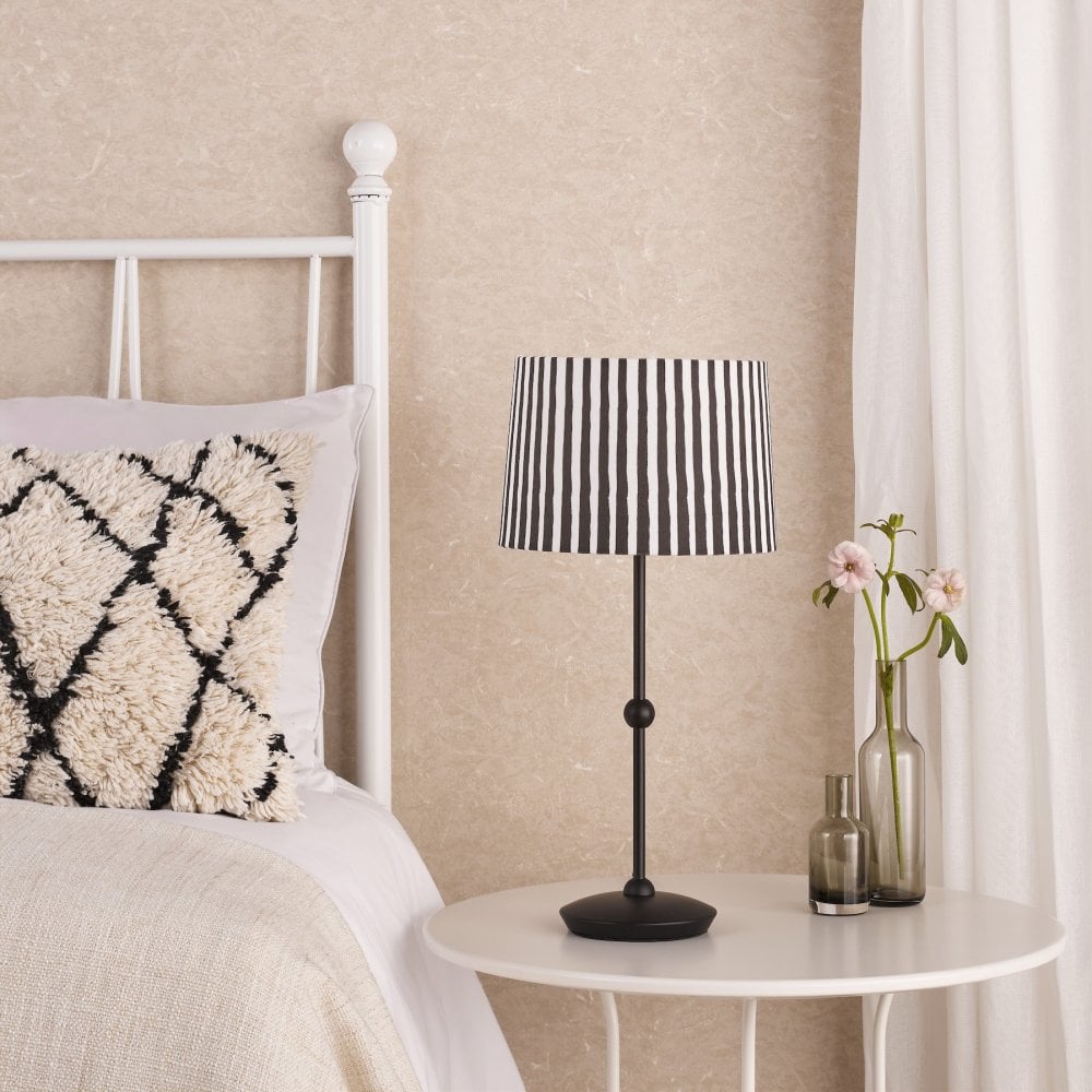 A black table lamp with a striped shade next to a bed.