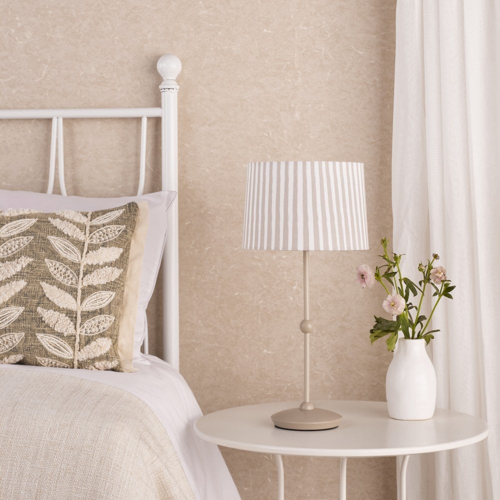 A pleated natural and off-white striped lampshade on a bedside table with a pillow and flowers.
