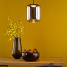 A modern pendant light illuminating a tabletop with a vase and bowl against a yellow wall.