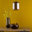 A modern pendant light illuminating a tabletop with a vase and bowl against a yellow wall.