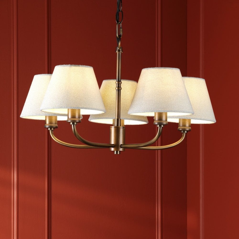 A five-light chandelier with white shades and an antique brass finish on a red paneled wall.