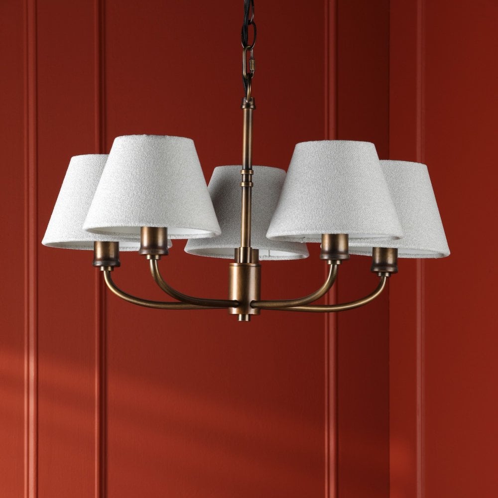 A five-light chandelier with grey shades and an antique brass finish on a red paneled wall.