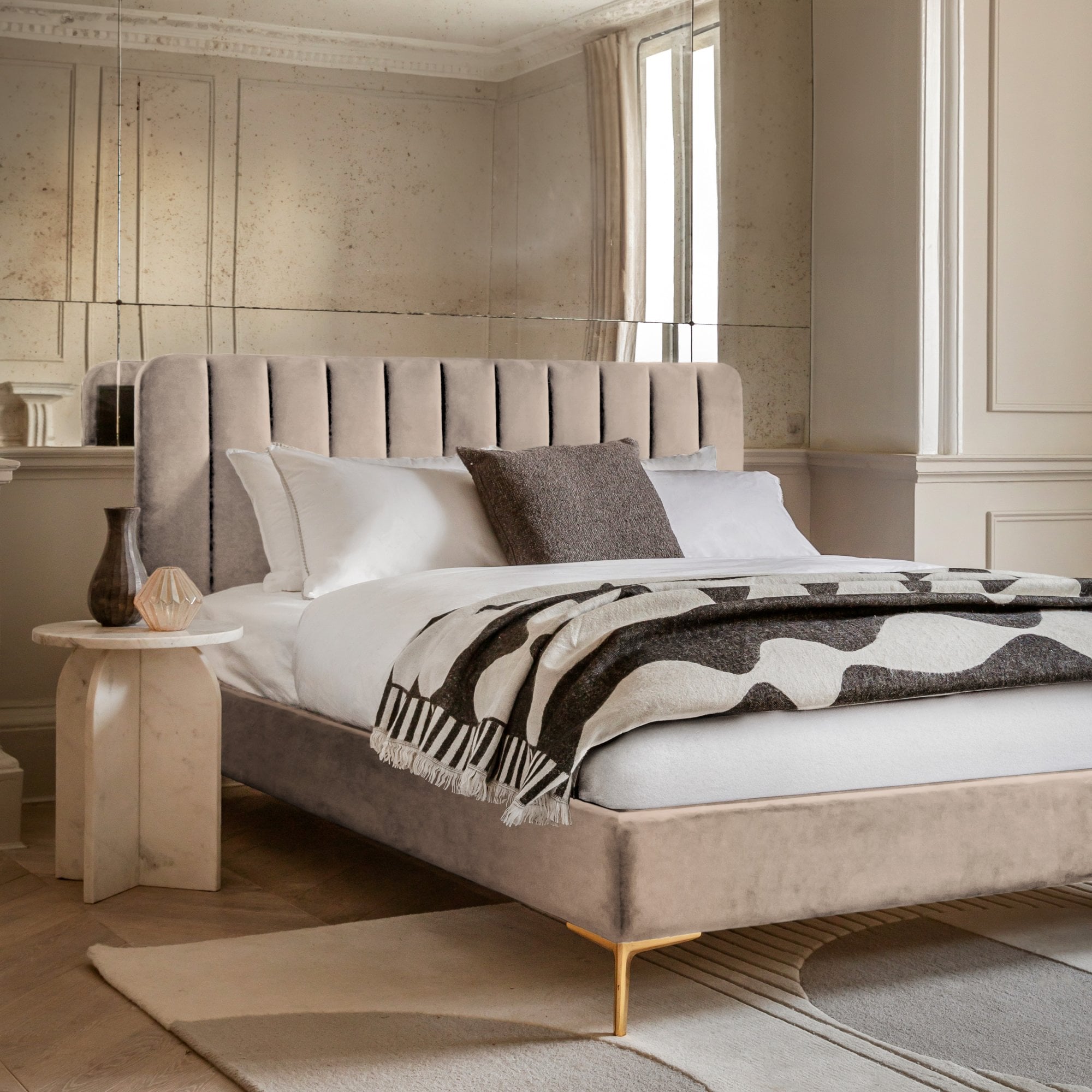 A beige upholstered bed with a channel-tufted headboard, gold legs, and a patterned throw.
