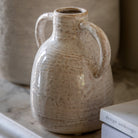 A close-up of a textured white ceramic vase with two small handles, on a marble shelf.