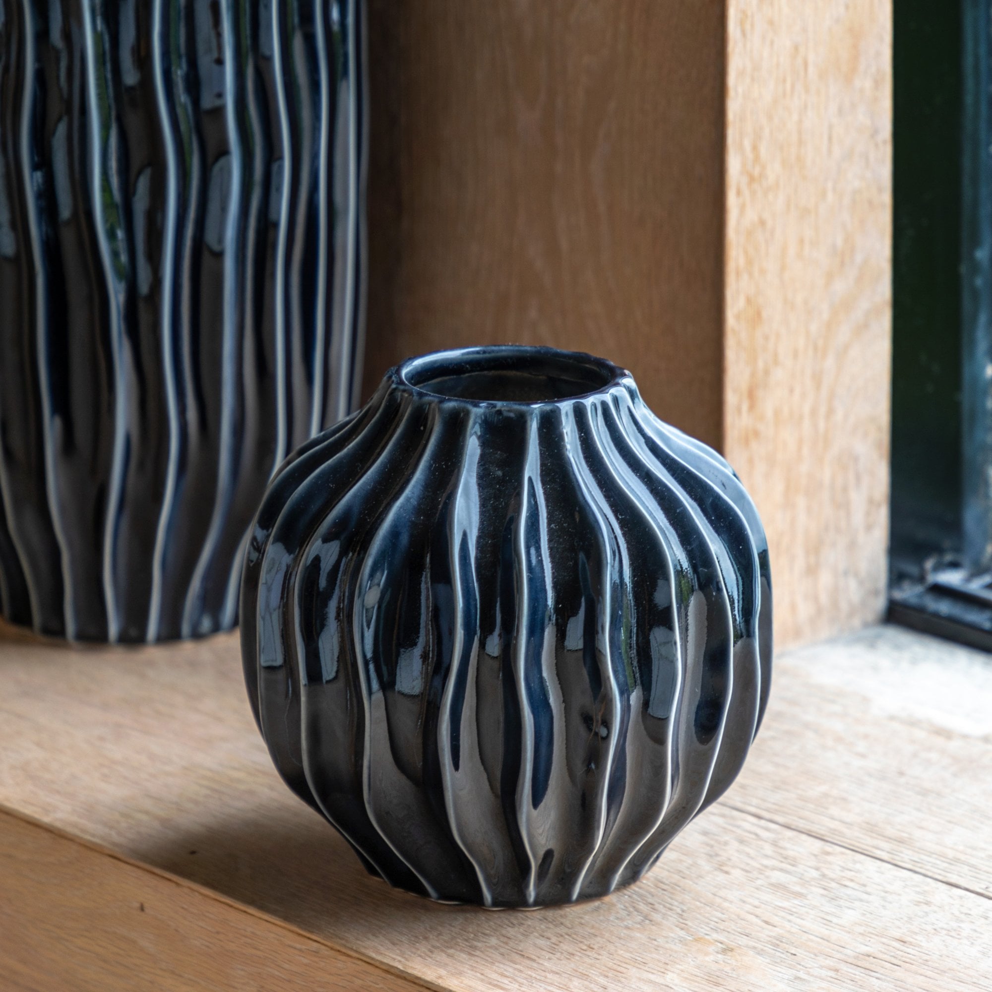 A short, black decorative vase with a wavy texture sits on a wooden shelf.