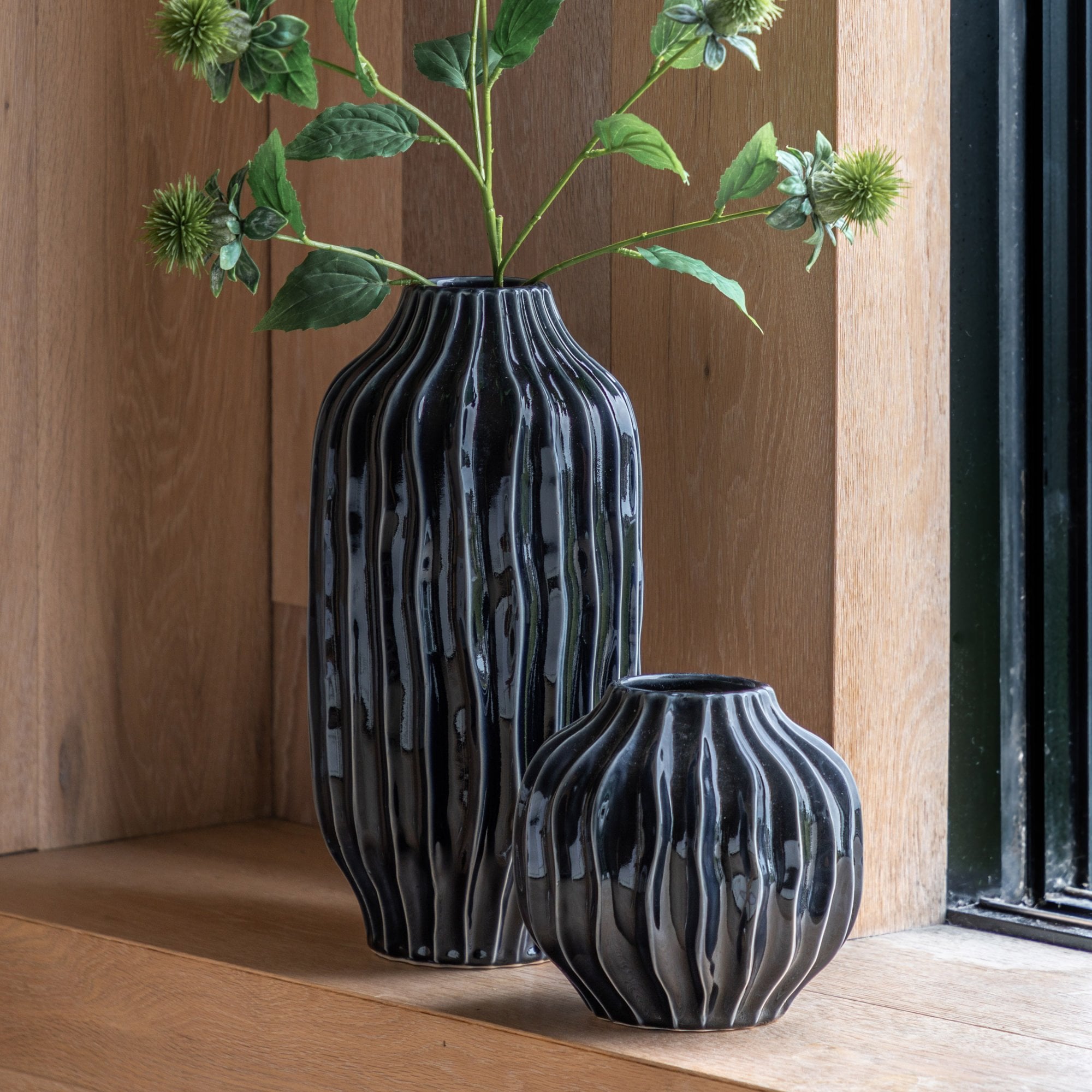 A tall and short black wavy textured vase on a wooden surface, with a plant in the tall vase.