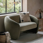 A modern, sage green velvet sofa with a patterned cushion sits by a large window.