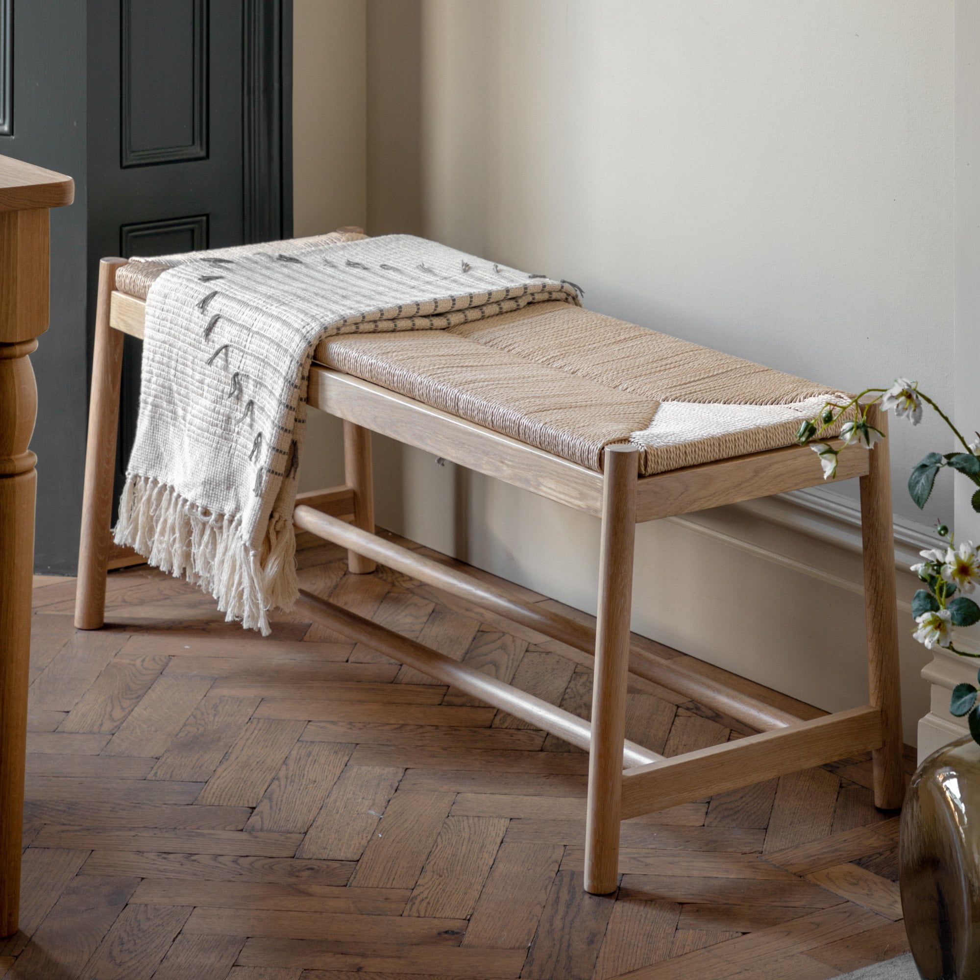 Light wooden bench with woven top and a fringed blanket draped over one end.