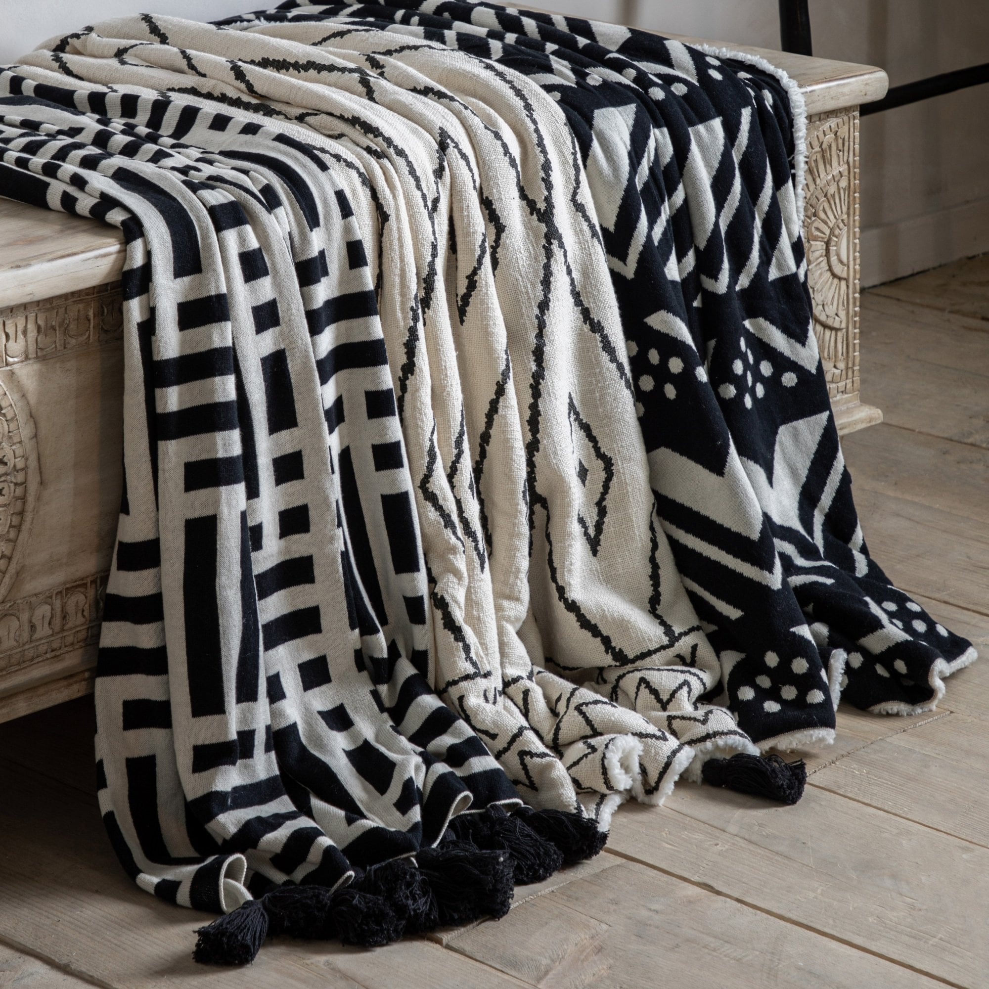 Three patterned throws draped over a rustic wooden bench, showcasing various black and white designs.